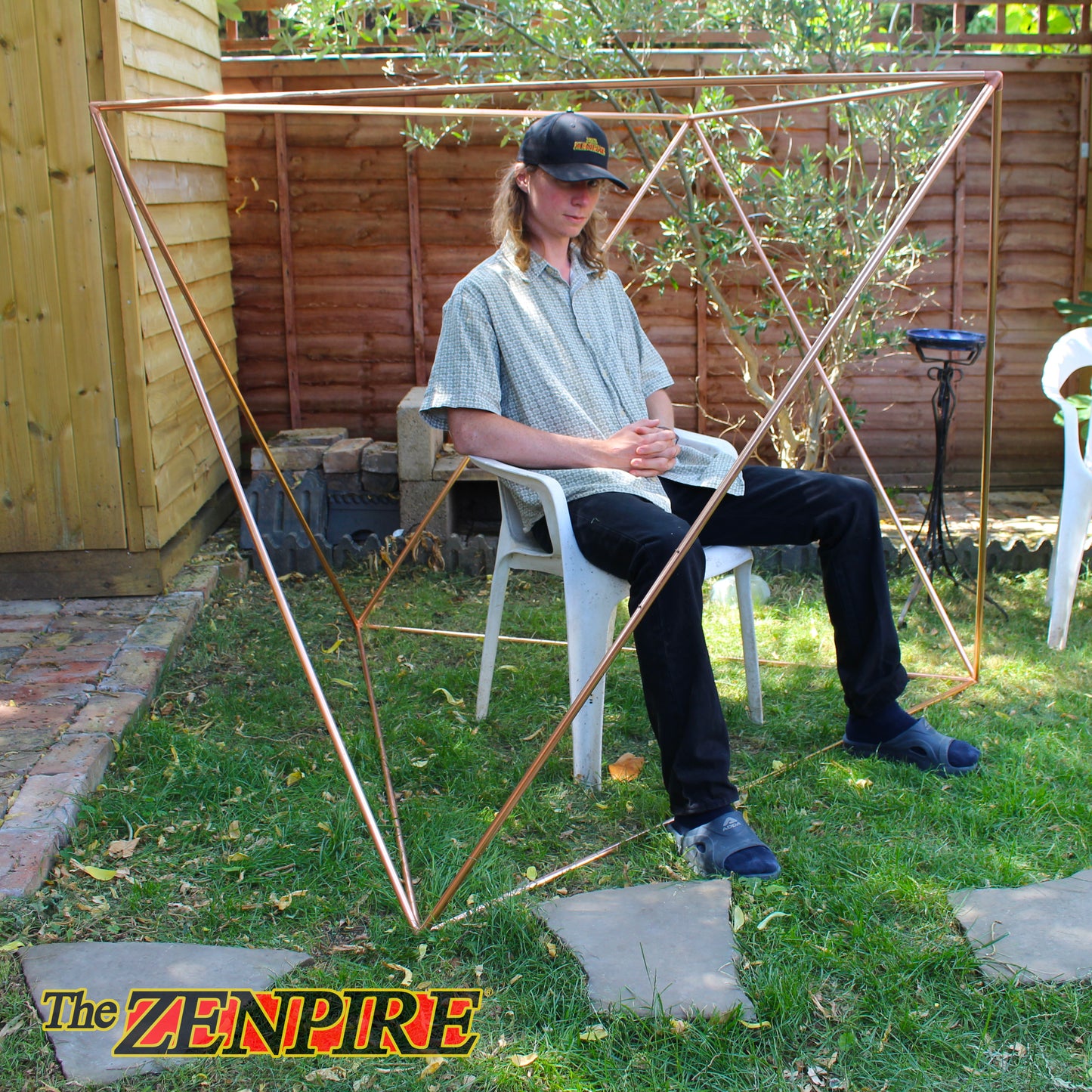 A man sits on a white plastic chair in a backyard, wearing a black cap and looking down. Surrounding him is a large, octahedron pyramid geometric frame made of copper pipes.