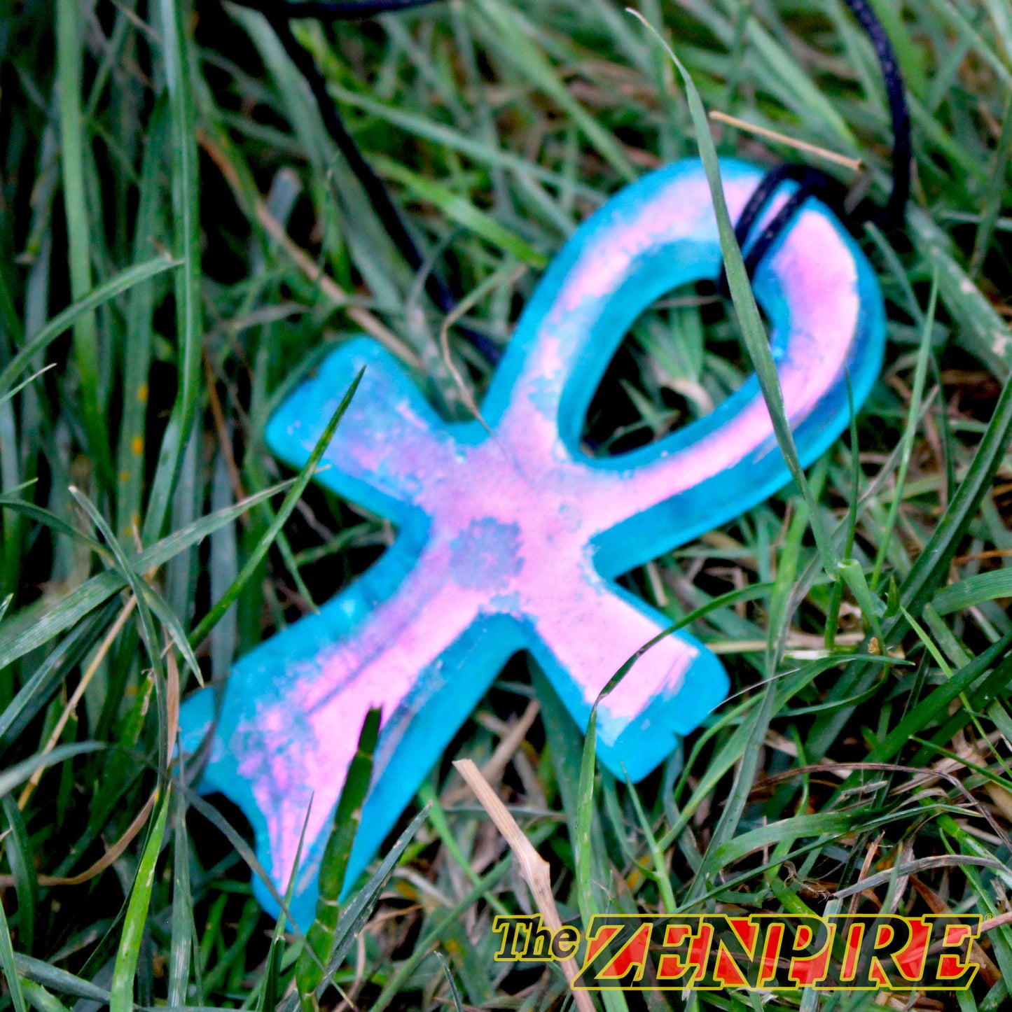 A blue and pink iridescent Atlantean Ankh pendant necklace lying on green grass, with a black cord visible at the top.