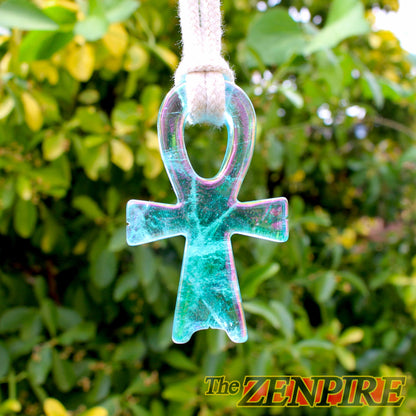A translucent iridescent clear quartz atlantean ankh symbol ornament hanging from a white cord, set against a blurred background of green foliage. The product features a blue-green base with pink and purple iridescent shiftsdue to its gold plating.