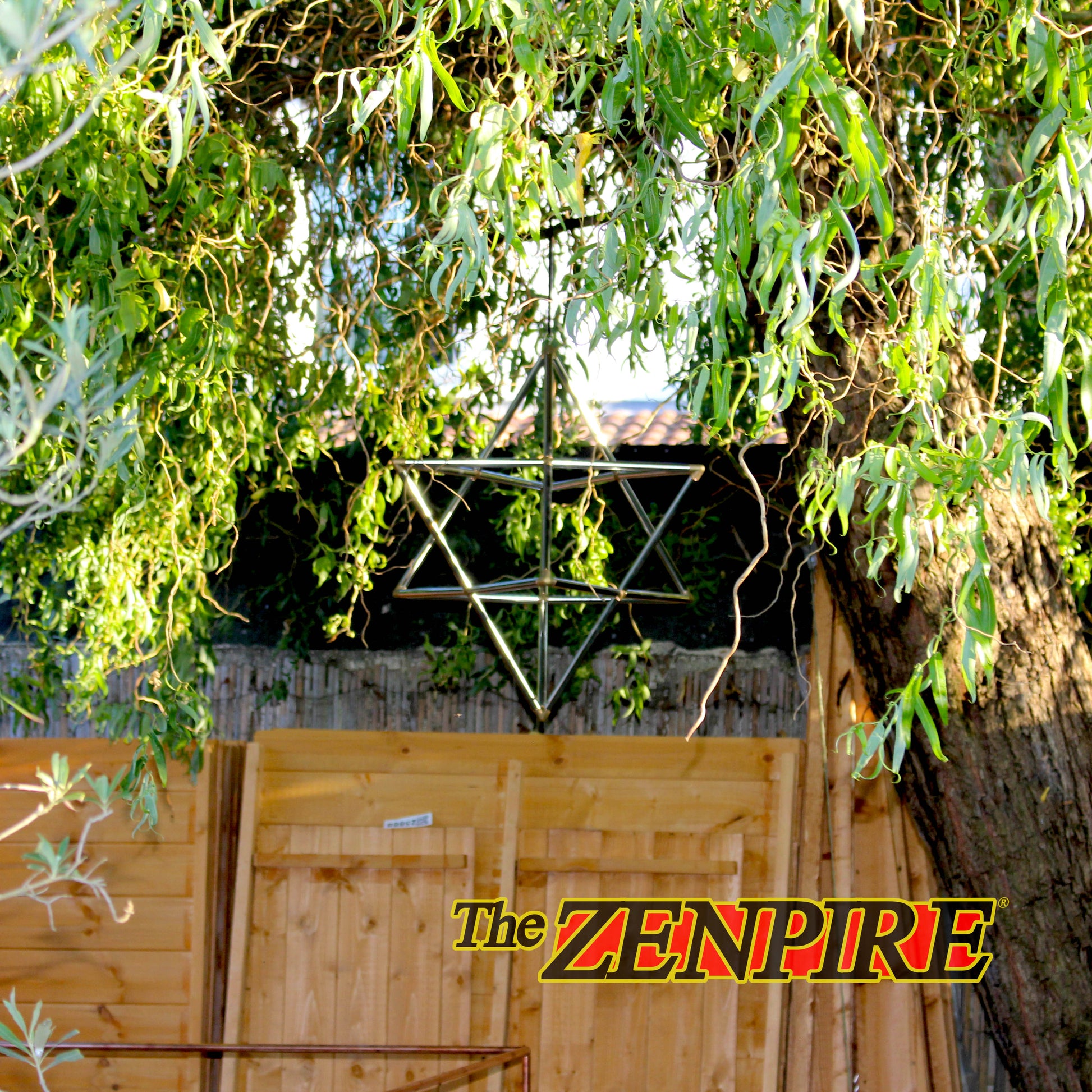A copper hanging merkaba ornament suspended outdoors beneath weeping willow branches in front of a wooden fence and tree trunk