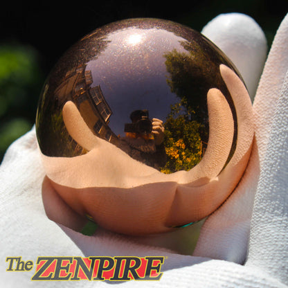A highly polished copper sphere held in a white-gloved hand, reflecting the photographer holding the camera and green trees in its surface. The Zenpire logo is visible in the bottom-left corner.