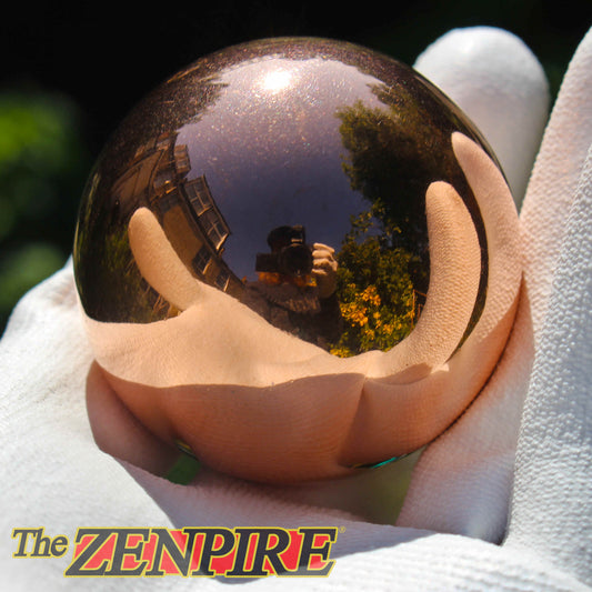 A highly polished copper sphere held in a white-gloved hand, reflecting the photographer holding the camera and green trees in its surface. The Zenpire logo is visible in the bottom-left corner.