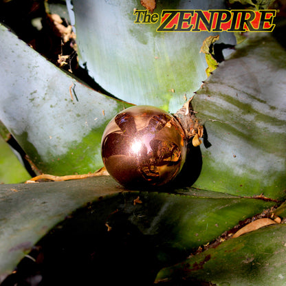 A highly reflective, metallic copper spherical ornament resting on the broad green leaves of a tropical plant, with 'The ZENPIRE' logo in the top right corner.