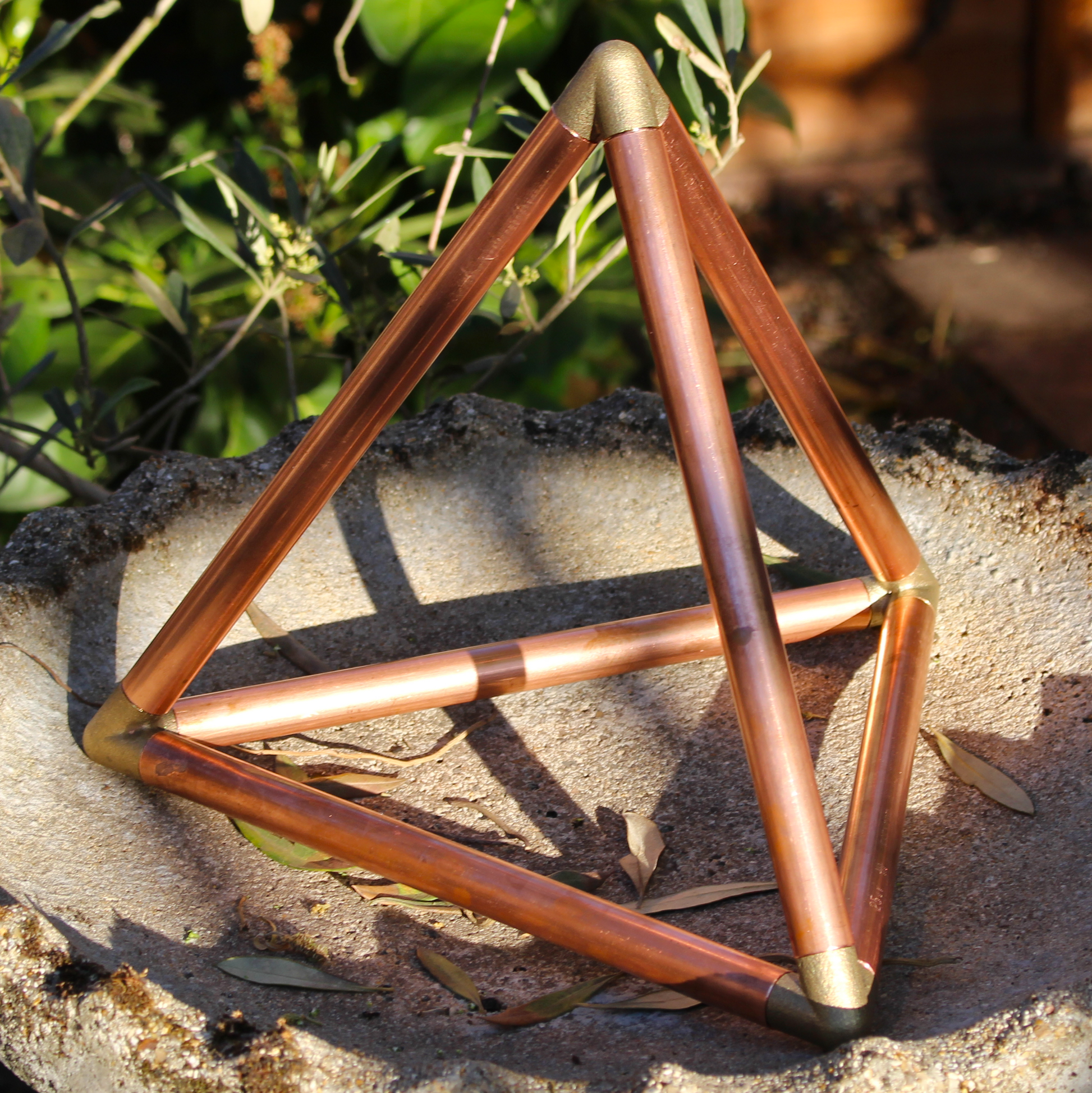 Copper tetrahedron sculpture on a stone surface with greenery in the background