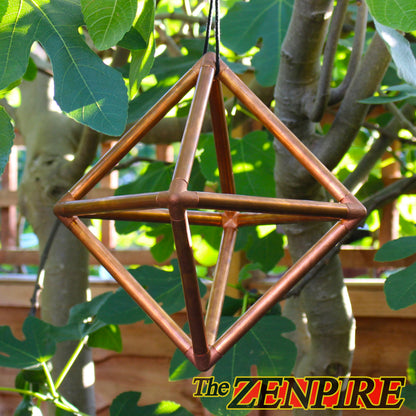 A geometric hanging ornament made of interlocking copper-colored metal rods, suspended outdoors among green leaves and tree branches.