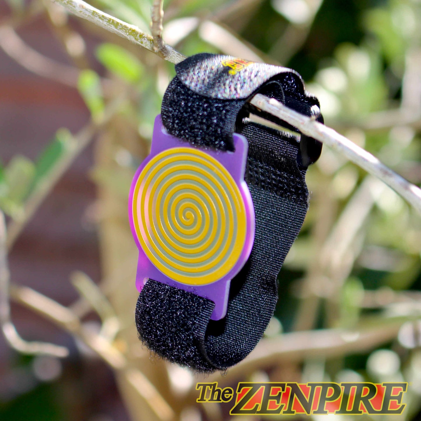 A black wristband with a purple and gold healing spiral, shown hanging from a tree branch outdoors