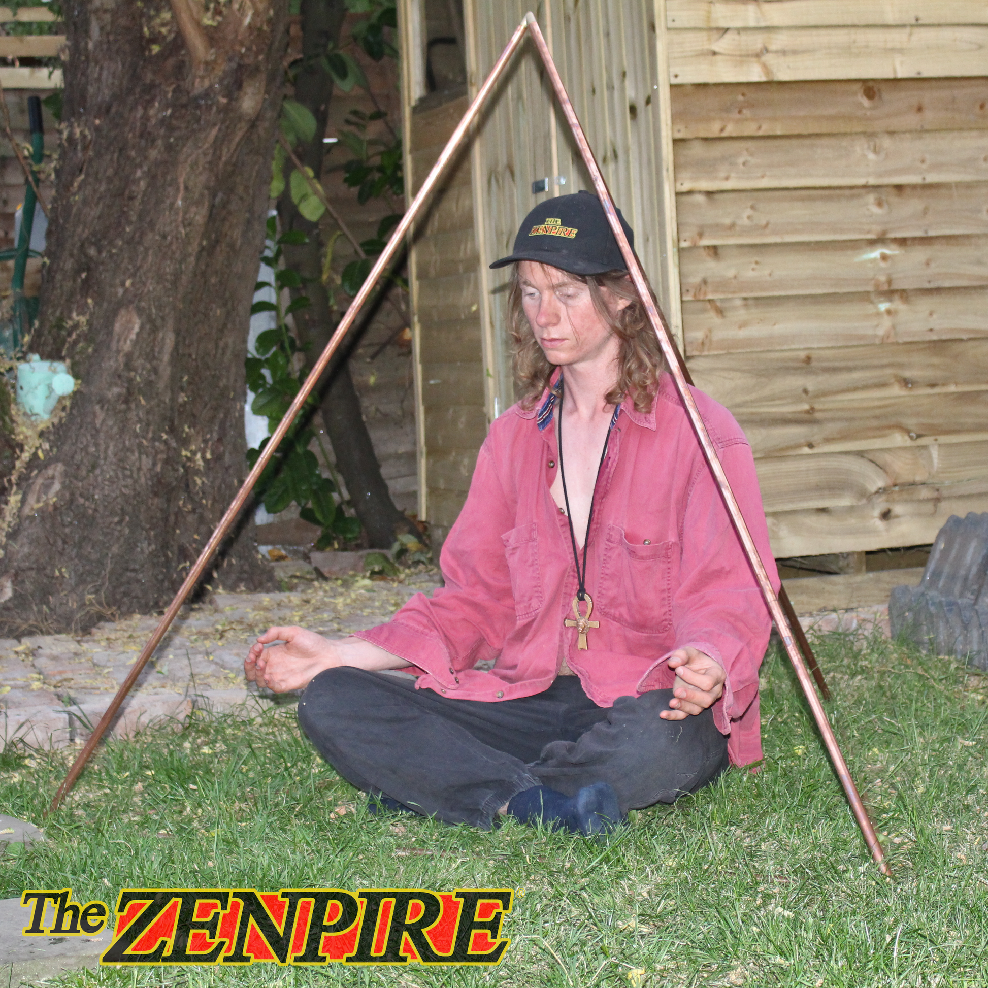 Person meditating outdoors with a copper pyramid kit structure and tree in the background, By The Zenpire