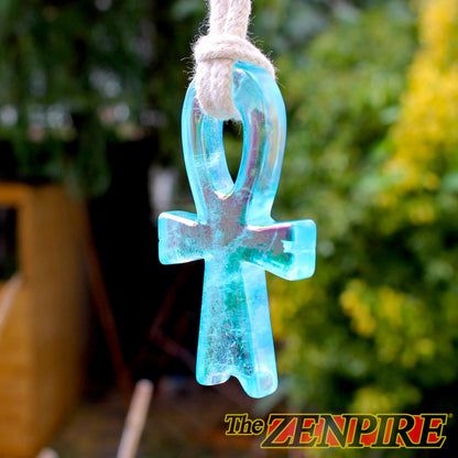 A translucent light blue quartz atlantis Ankh pendant hanging from a white rope, with a blurred garden background and 'The ZENPIRE' logo in the bottom right corner.
