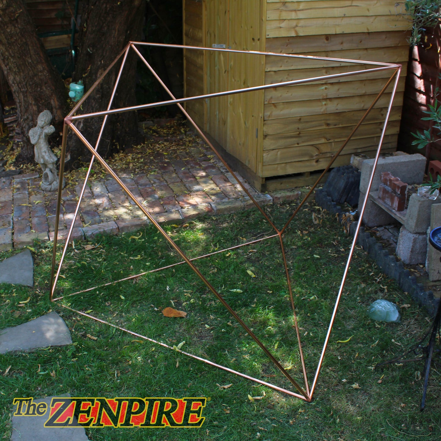 A geometric octahedron wireframe structure made of copper pipes, set outdoors on a grassy lawn, used for meditation.