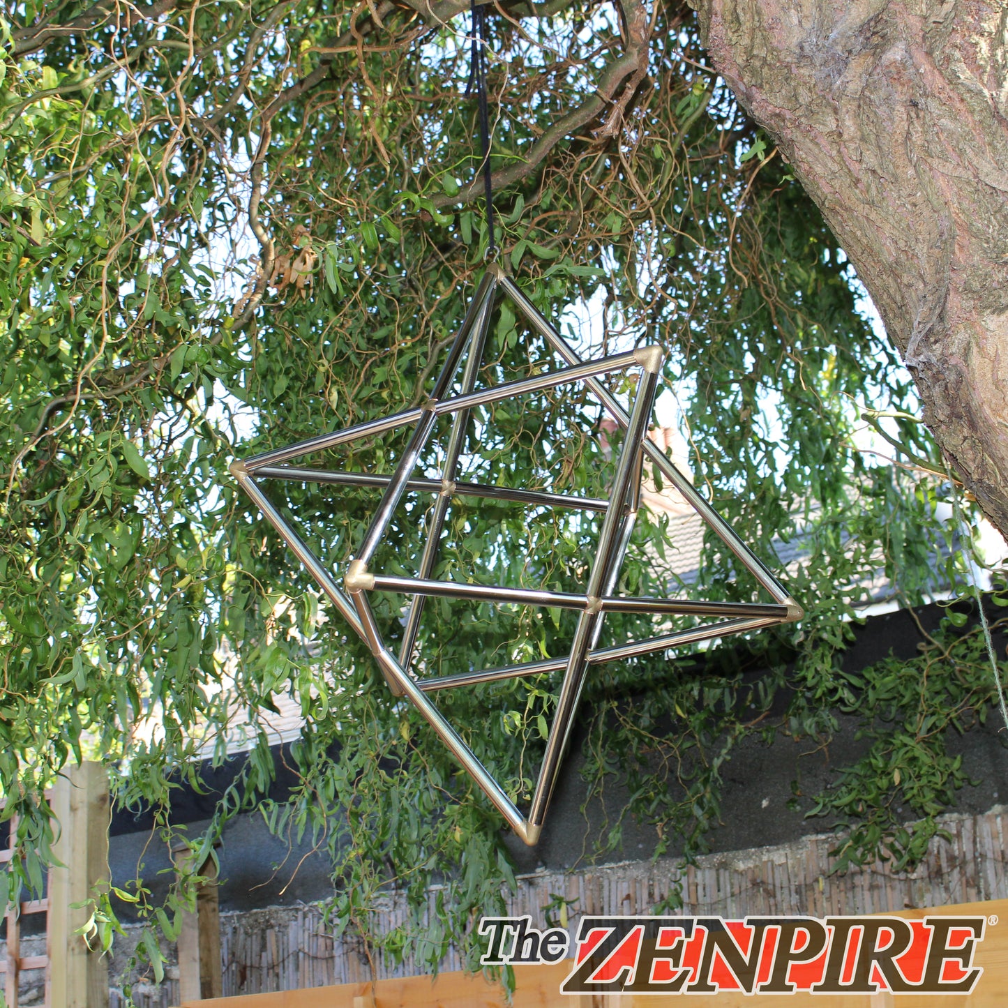 A metal geometric hanging ornament made of intersecting silver-toned rods, suspended outdoors from a tree branch with green leaves and a fence in the background. The brand name 'The ZENPIRE' is visible as an overlay at the bottom right.